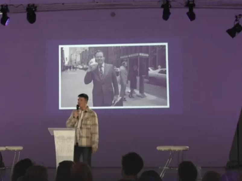 Thijs op het podium met een microfoon, voor een projectiescherm, met publiek op de voorgrond